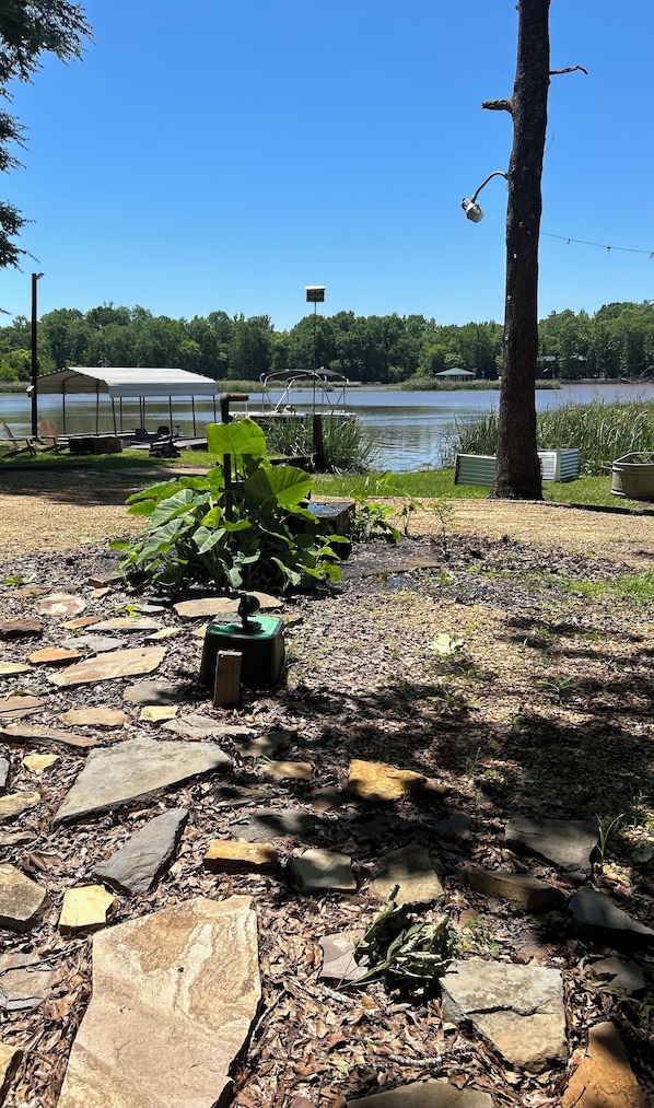 A lakeside area with a covered dock, stone pathway, and a large tree.