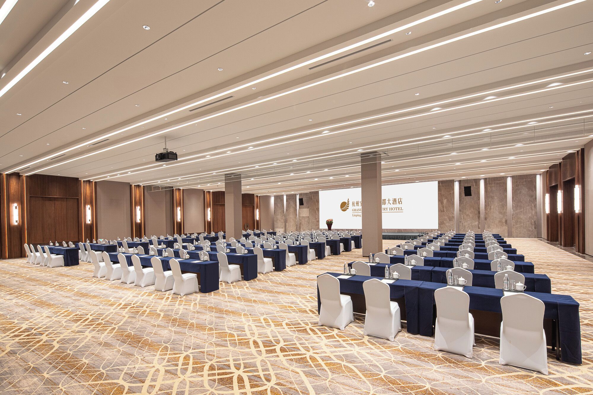 Una sala conferenze con file di tavoli e sedie, un proiettore e uno schermo con testo.