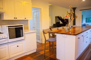 A kitchen with white cabinetry, a wooden countertop island, and a piano in the background. - The Farmhouse at Levenvale Farm (Bellingen)