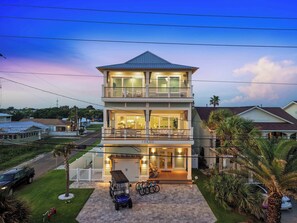 A two-story residential building with a balcony on the upper floor, a garage at the front, and a driveway with a golf cart and bicycles. - Casa Elcoqui | LUX Gulf Views, Pool + Spa, Sleeps 16 (Panama City Beach)
