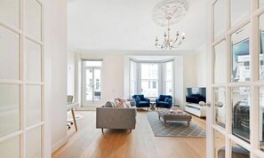 A spacious living room with a large window, a chandelier, a sofa, two armchairs, and a television. - Luxury Refurbished 3BR With Patio Kensington (London)