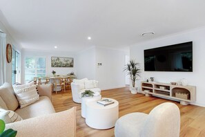 Ein modernes Wohnzimmer mit Flachbildfernseher, weißen Sofas, einem Couchtisch und einem Essbereich mit Tisch und Stühlen.