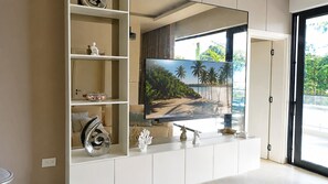Ein modernes Wohnzimmer mit großem Flachbildfernseher an einer weißen Unterhaltungseinheit, einem Glastisch und dekorativen Elementen, darunter eine Muschel und eine Pferdefigur.