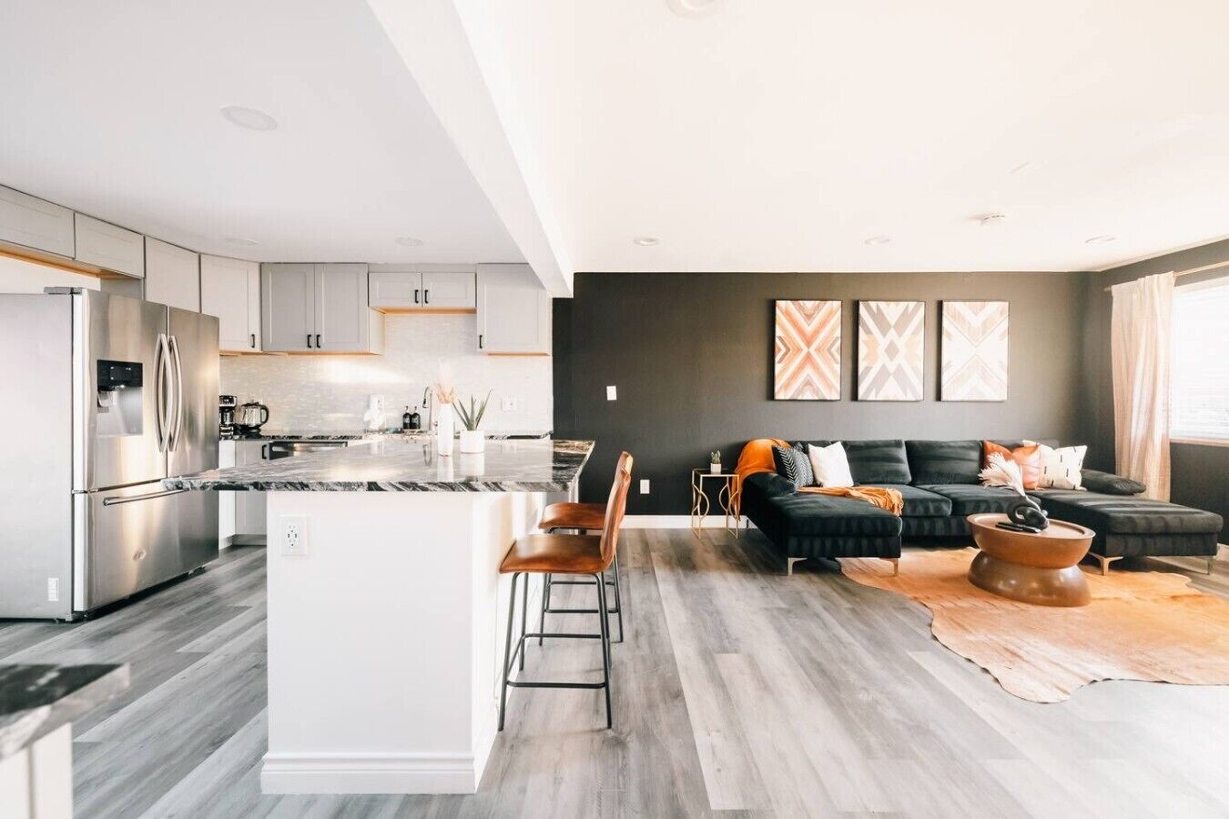 A modern kitchen with a central island, a dining area with a black sofa and chairs, and a living room with a large black sofa and decorative pillows.