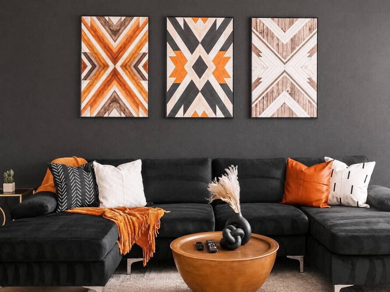 Un salon moderne avec un canapé noir, des coussins orange et blancs, une table basse en bois et trois œuvres d’art abstraites accrochées au mur.