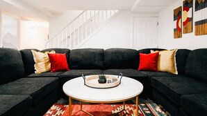 A modern living room with a sectional sofa, a round coffee table, and a staircase in the background. - 5 BR | Hot Tub•Game Room•Fire Pit•Big Backyard (Westminster)