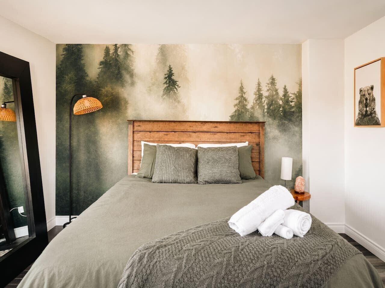 A bedroom with a bed, a headboard, a wall mural, a lamp, and a framed picture.