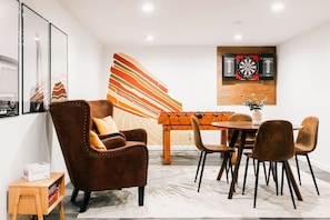 Ein modernes Zimmer mit einem braunen Ledersessel, einem runden Esstisch mit Stühlen, einem Dartspiel und einem farbenfrohen abstrakten Gemälde an der Wand.
