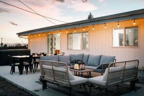 An outdoor seating area with a sofa, chairs, and a table, connected to a building with large windows and string lights. - 5 BR | Hot Tub•Game Room•Fire Pit•Big Backyard (Westminster)