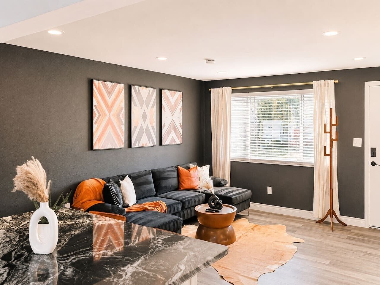A modern living room with a sectional sofa, a coffee table, and a rug. There is a window with curtains and a door to the right.