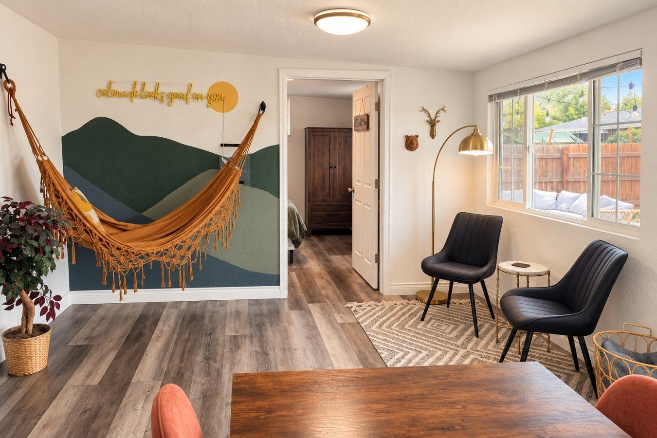 A room with a hammock, a dining table, two chairs, a lamp, and a wall with a deer head decoration.