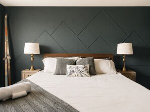 A neatly made bed with pillows and a blanket, two bedside lamps, and a dark wall with a geometric pattern. - 5 BR | Hot Tub•Game Room•Fire Pit•Big Backyard (Westminster)