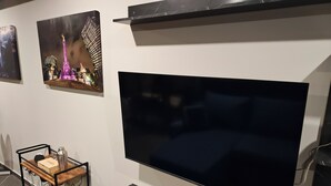 Una sala moderna con un televisor de pantalla plana grande montado en la pared, una mesa de centro de madera con una botella y una caja, y un estante pequeño debajo del televisor.