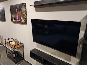 Ein modernes Wohnzimmer mit einem großen Flachbildfernseher an der Wand, einem Couchtisch aus Holz mit einer Flasche und einer Schachtel, sowie einem kleinen Schrank darunter.