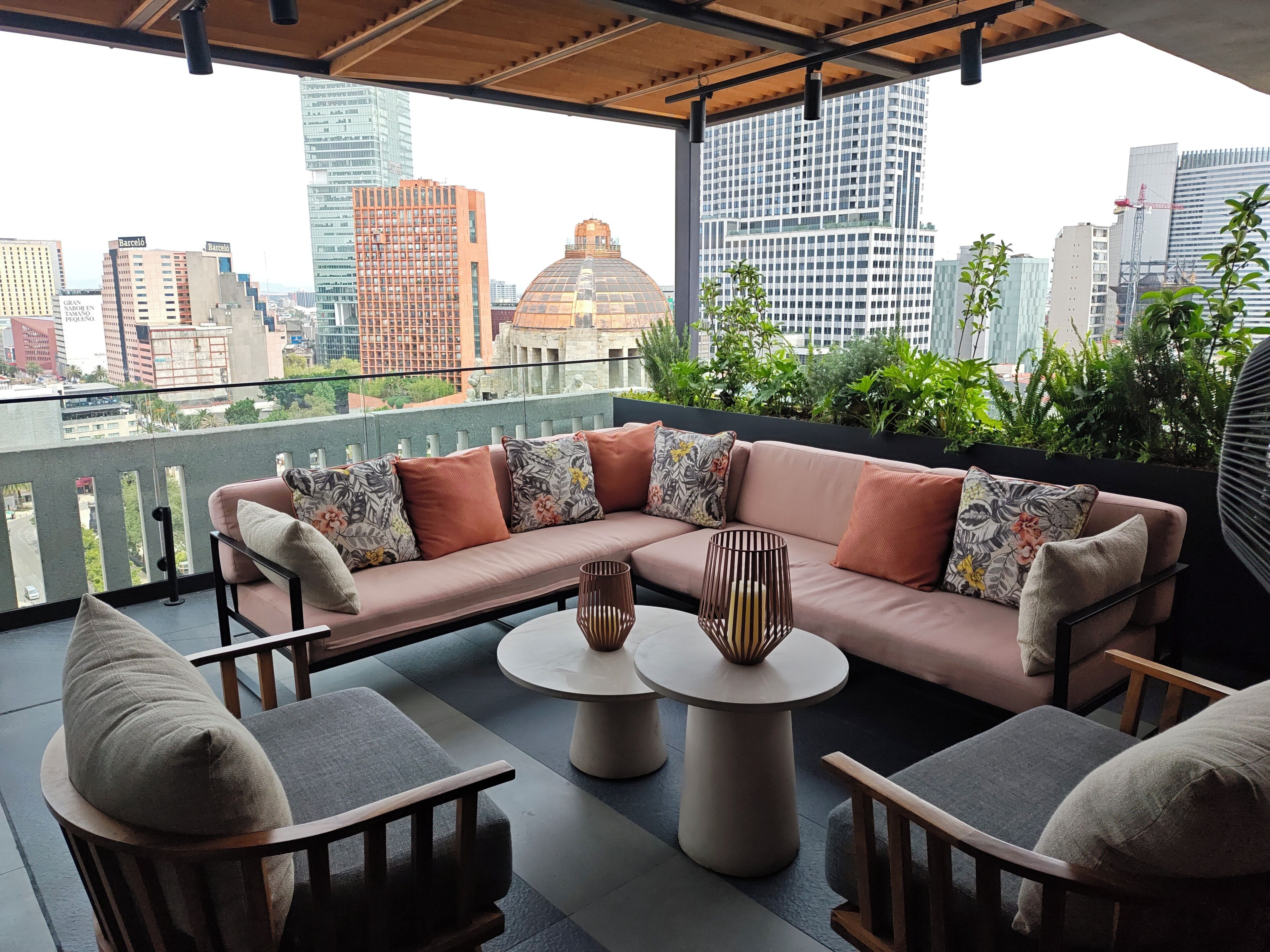 A balcony with a sofa, chairs, and a table, overlooking a cityscape with buildings and greenery.