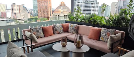 A balcony with a sofa, chairs, and a table, overlooking a cityscape with buildings and greenery.