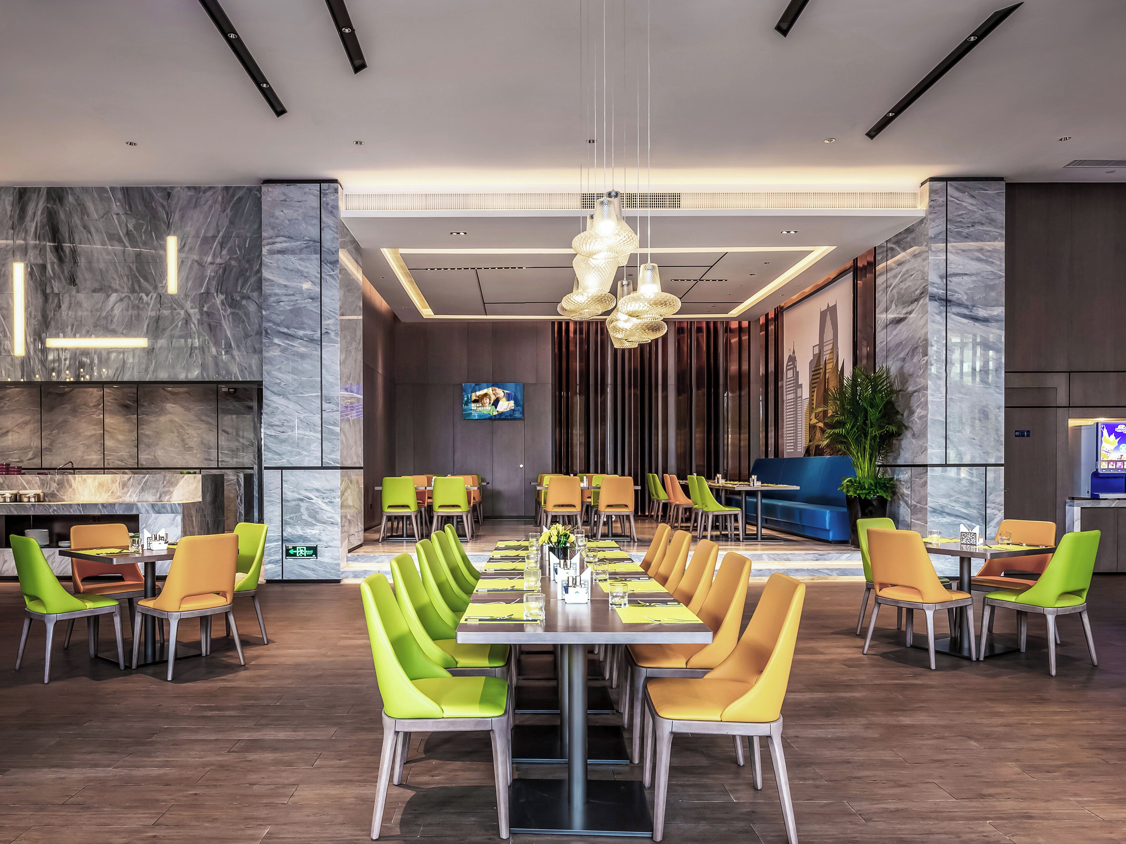 Un espace de repas moderne avec une longue table et de nombreuses chaises, un écran de télévision et un canapé bleu en arrière-plan.