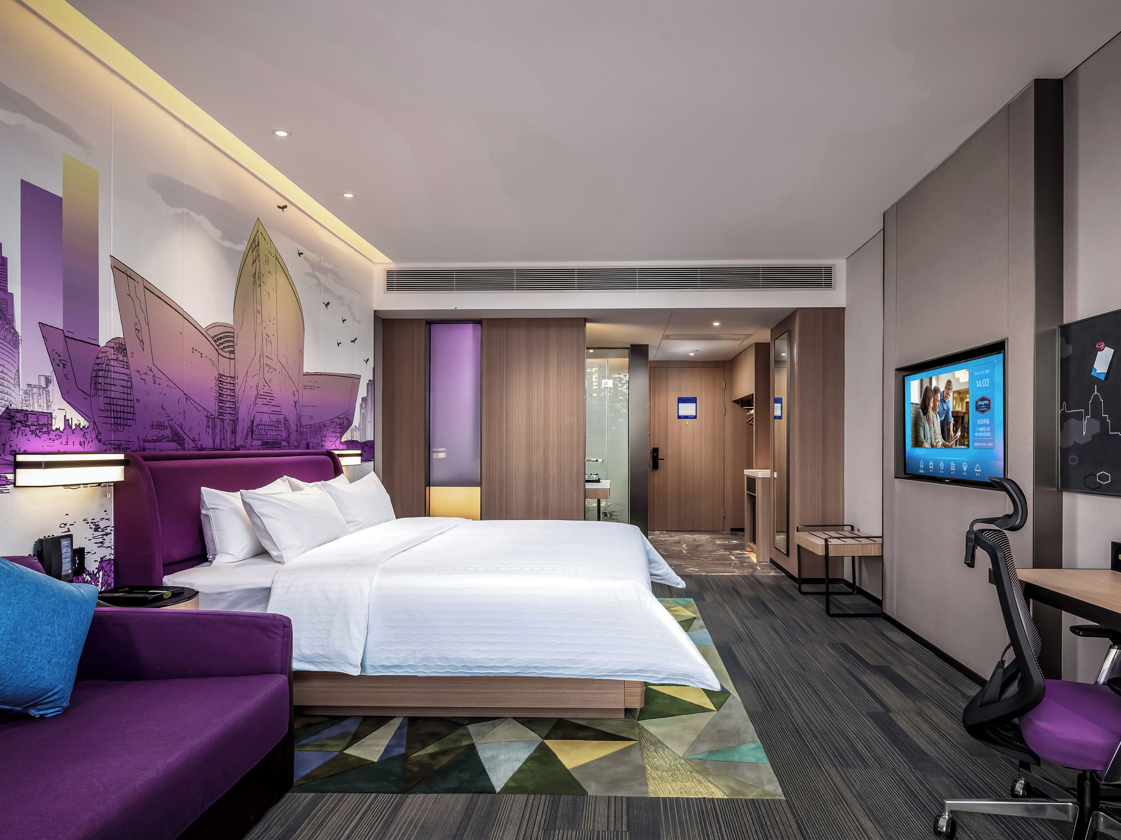 A modern hotel room with a bed, a desk, a chair, a TV, and a cityscape mural on the wall.