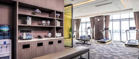 A hotel room with a bench, water dispenser, shelves with books and decorative items, exercise equipment, and large windows.