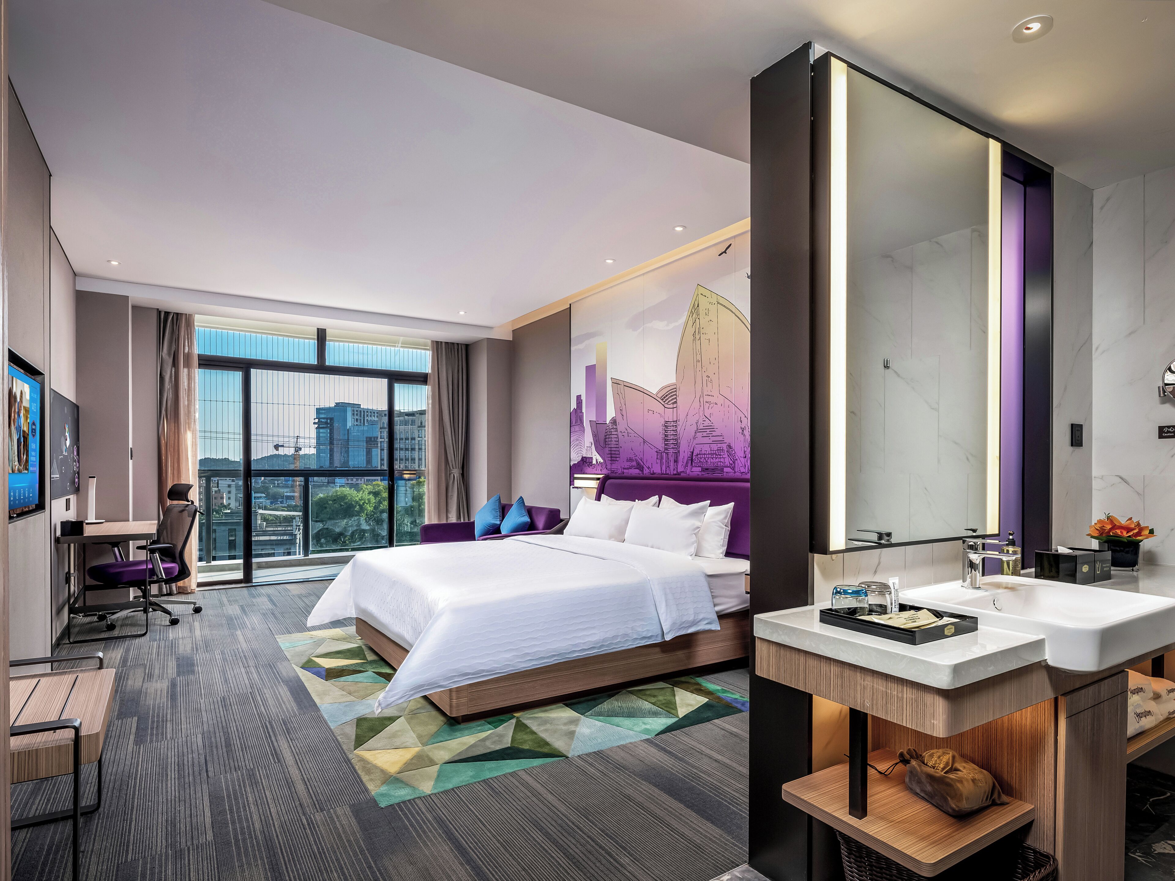 Una habitación de hotel moderna con una cama grande, un escritorio, una silla, un baño con espejo y vistas a la ciudad.