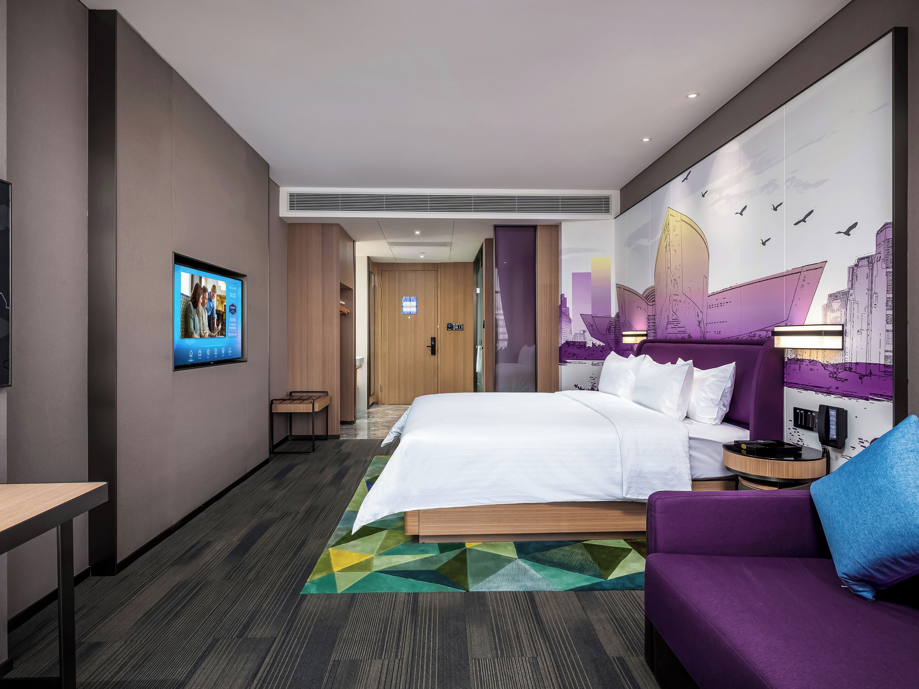 A modern hotel room with a bed, a seating area, a television, and a cityscape mural.