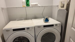 Two Whirlpool front-loading washing machines in a laundry room with a metal rack above and a door to the right. - New 4-Bedroom Home | Modern Comfort in NE Calgary—Beautiful and Affordable (Calgary)