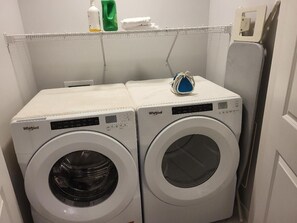 Two Whirlpool front-loading washing machines in a laundry room with a metal rack above and a door to the right. - New 4-Bedroom Home | Modern Comfort in NE Calgary—Beautiful and Affordable (Calgary)