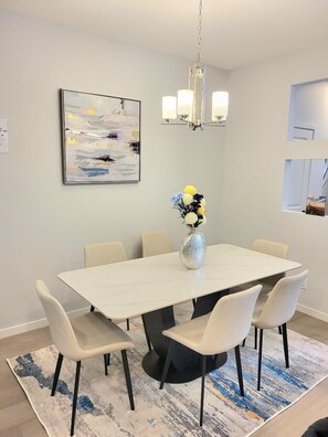 A dining area with a white table, six chairs, a vase with flowers, a chandelier, and a painting on the wall. - New 4-Bedroom Home | Modern Comfort in NE Calgary—Beautiful and Affordable (Calgary)