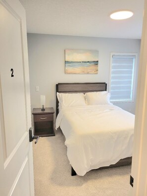 A bedroom with a bed, a nightstand, a painting, and a window with blinds. - New 4-Bedroom Home | Modern Comfort in NE Calgary—Beautiful and Affordable (Calgary)