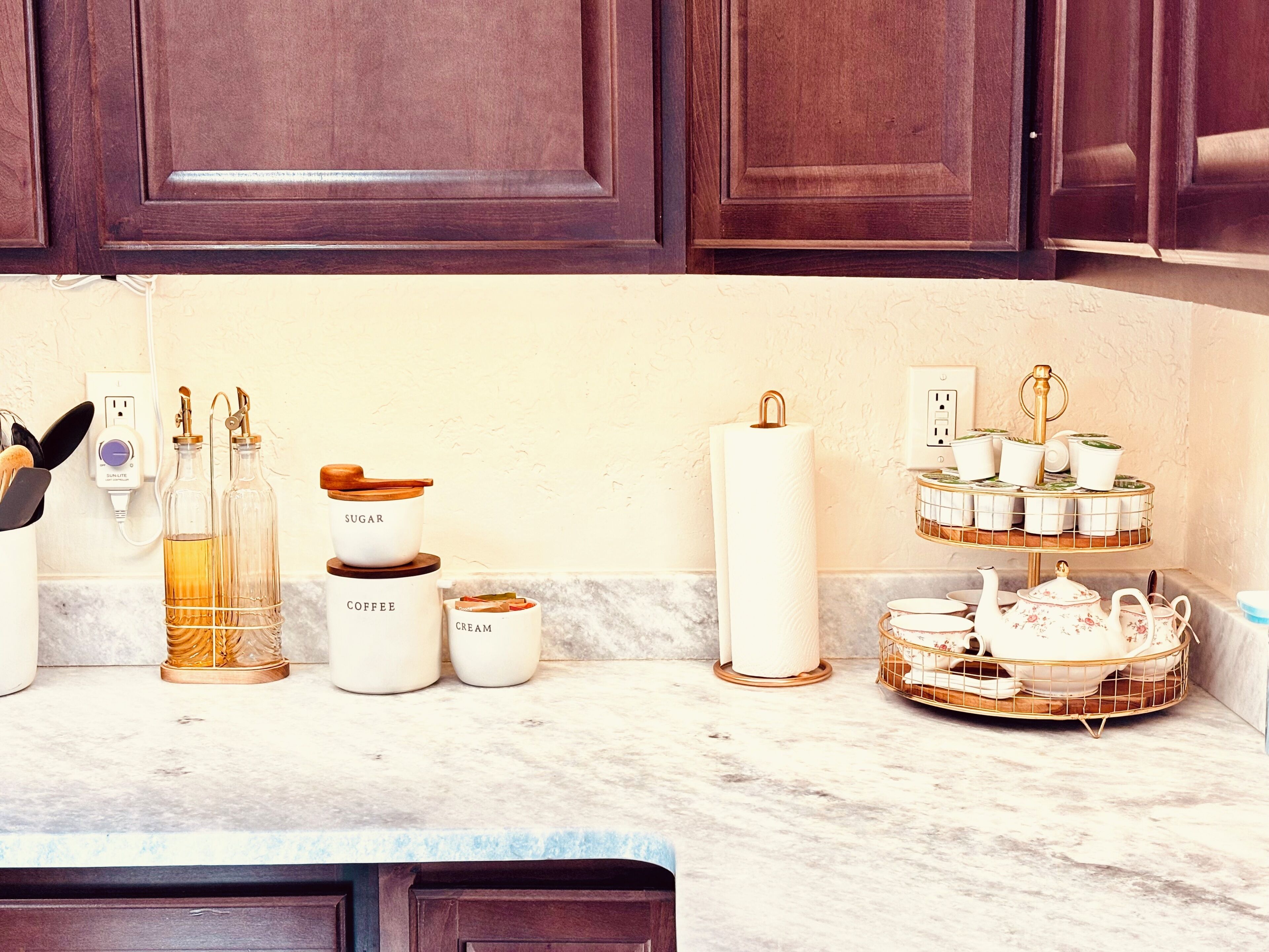 Una encimera de cocina con diversos objetos, incluyendo botellas con las etiquetas 'SUGAR', 'COFEE' y 'CREAM', un rollo de servilletas de papel blanco, y un frutero escalonado con teteras y tazas.