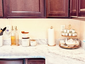 Una encimera de cocina con diversos objetos, incluyendo botellas con las etiquetas 'SUGAR', 'COFEE' y 'CREAM', un rollo de servilletas de papel blanco, y un frutero escalonado con teteras y tazas.