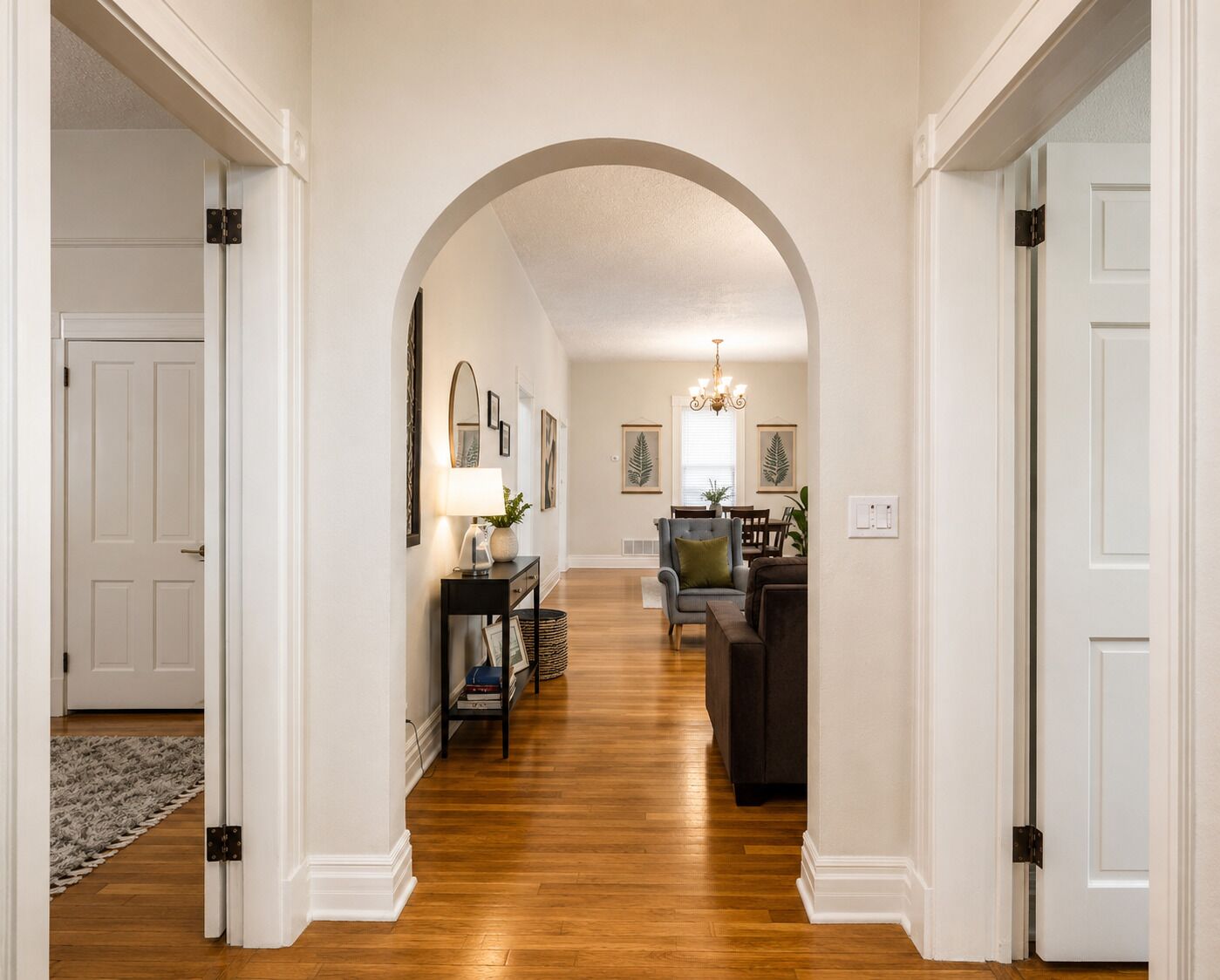 A hallway with a wooden floor, white walls, and an arched ceiling. There is a dining area with a table and chairs in the distance.