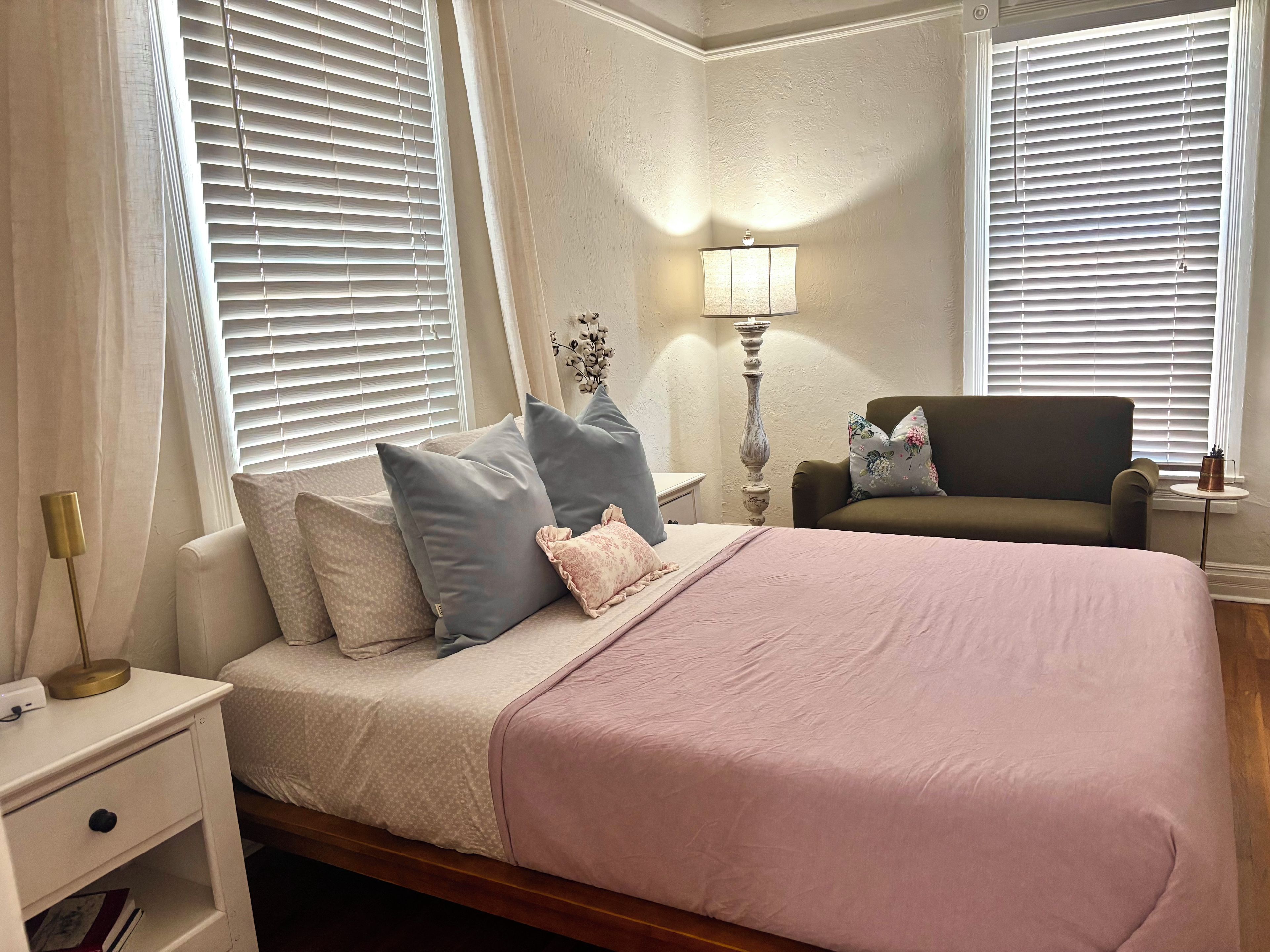 A bedroom with a bed, nightstand, lamp, chair, and window blinds.