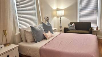 A bedroom with a bed, nightstand, lamp, chair, and window blinds.