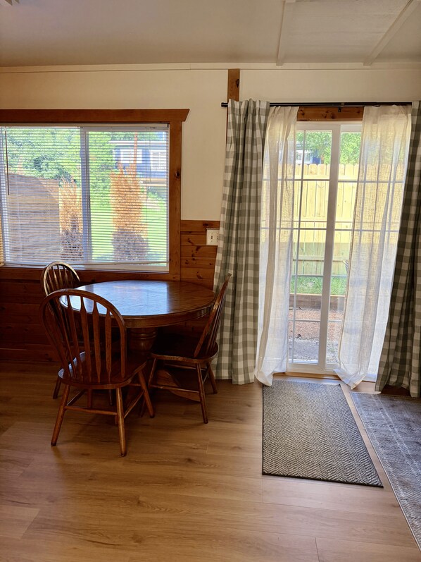 Een eethoek met een houten tafel en stoelen, een raam met jaloezieën en uitzicht op een houten schutting.