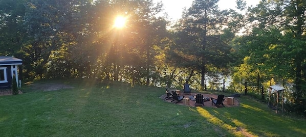 A grassy outdoor area with a barbecue setup and a small structure, likely a gazebo, in a wooded setting.