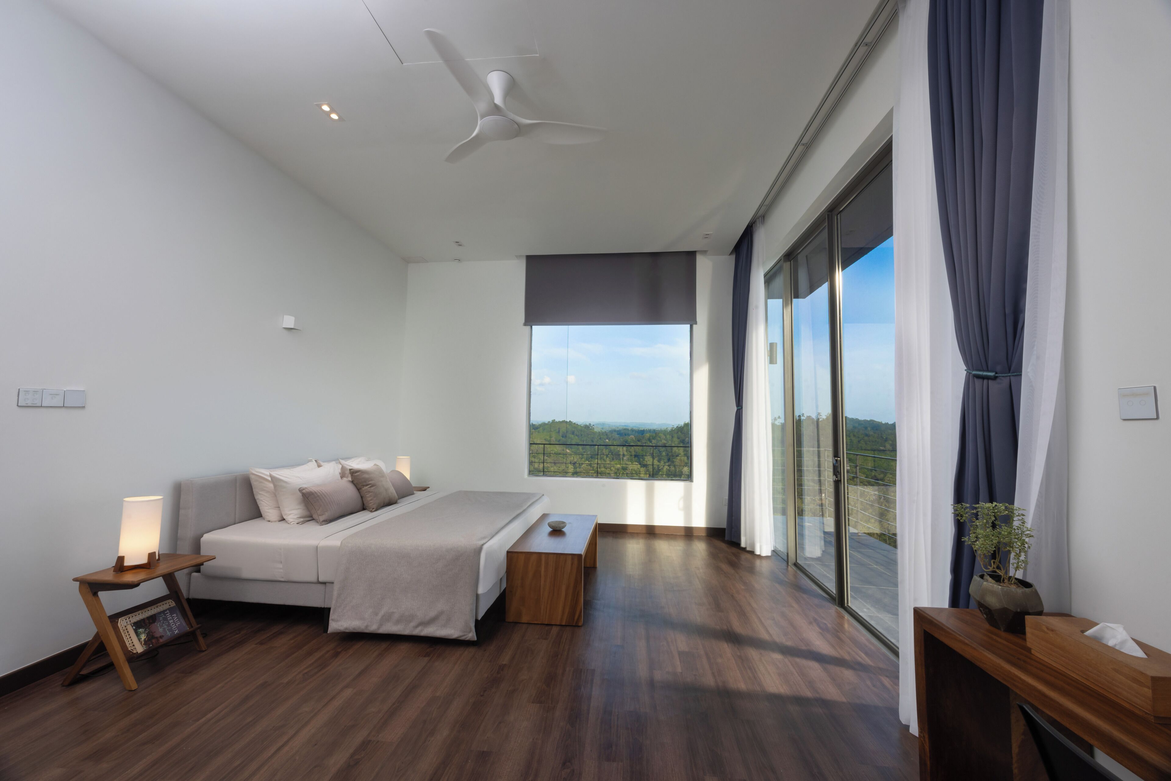 A modern bedroom with a large bed, a bedside table with a lamp, a wooden nightstand, and a view of the outdoors through a large window.