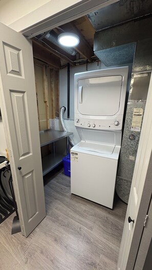 A compact laundry room with a front-loading washing machine, a small sink, and a blue trash bin.