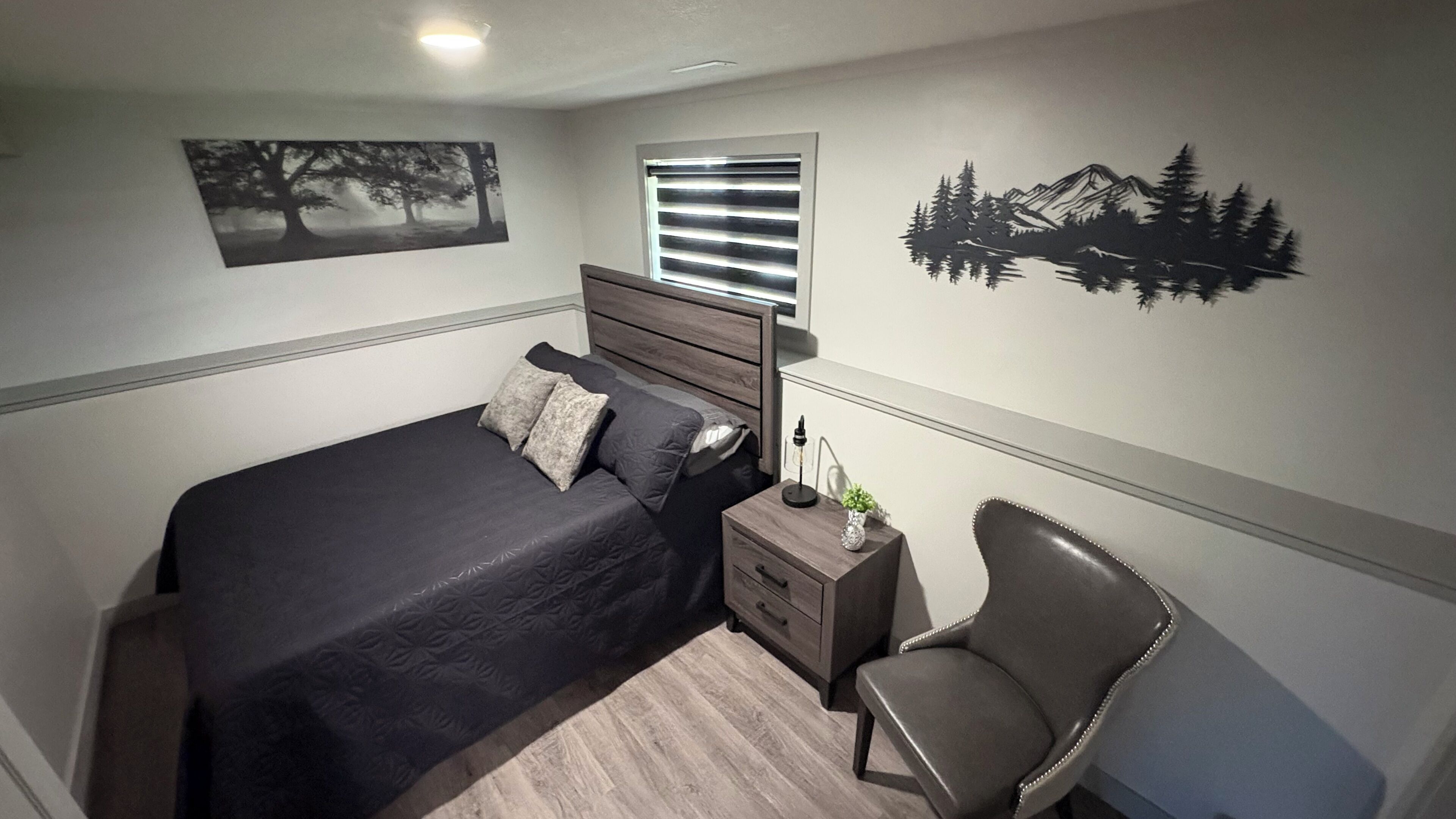 Een slaapkamer met een bed, nachtkastje, stoel, raam met jaloezieën en wanddecoratie.