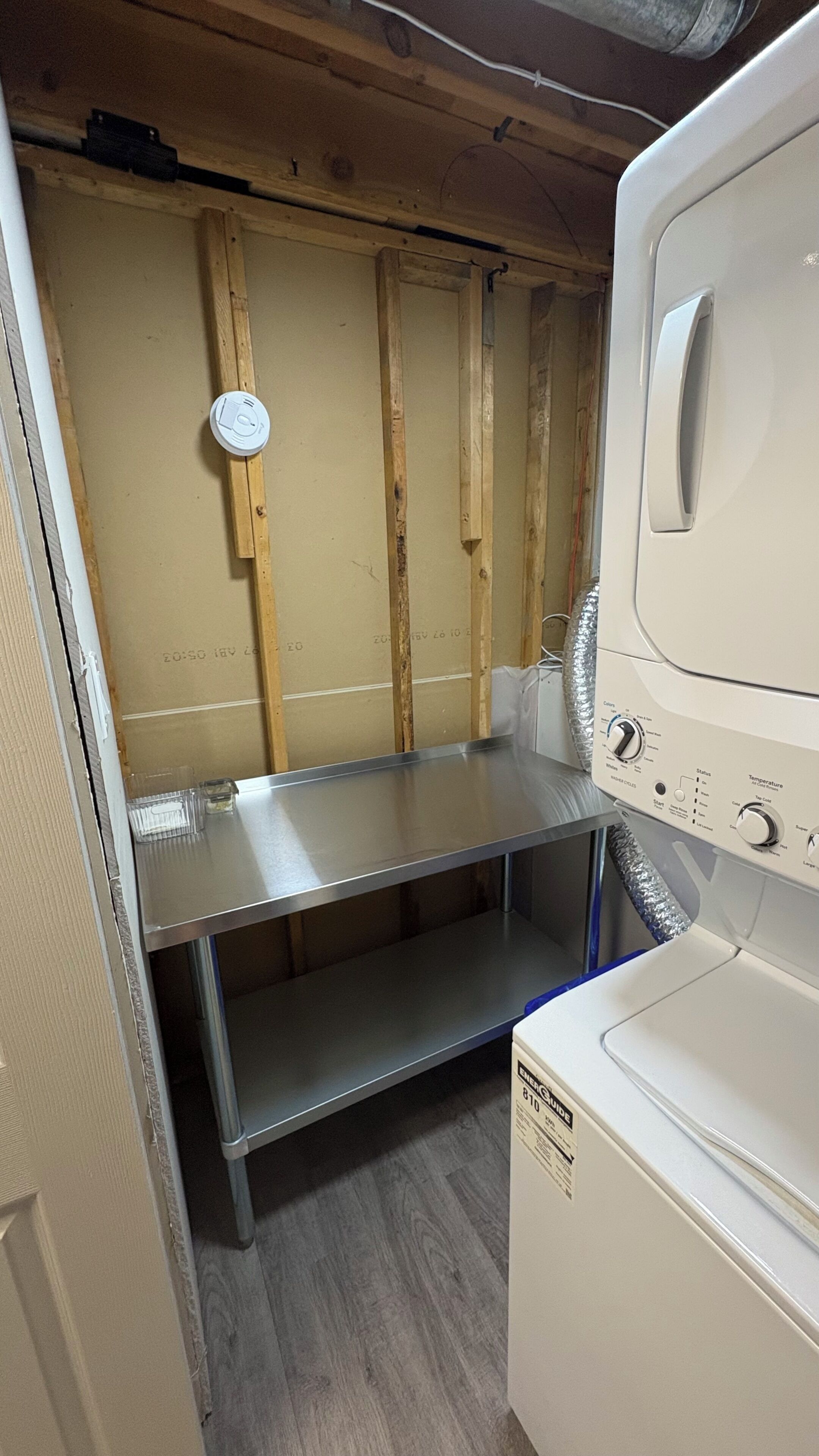Une buanderie équipée d’un lave-linge et d’un sèche-linge superposés, d’une étagère en métal et de montants muraux en bois.