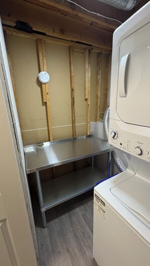 A laundry room with a stacked washer and dryer, a metal shelf, and wooden wall studs. - Stylish 2 bed 1 bath basement suite (Grande Prairie)