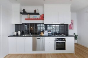 A modern kitchen with white cabinetry, black countertops, a dishwasher, a stove, and a sink. There are two shelves above the sink with various items. - Laidback Lygon A Two Storey Inner City Oasis (Brunswick East)