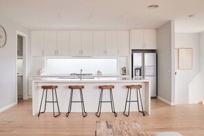 A modern kitchen with white cabinetry, a central island with stools, a refrigerator, and a window overlooking the interior. - Harbour Horizons Balcony BBQs and Calm Water Views (Clifton Springs)