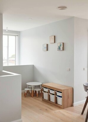Une pièce moderne et minimaliste avec un mur blanc, un meuble en bois doté de tiroirs, une petite table blanche avec des chaises et une fenêtre laissant entrer la lumière naturelle.