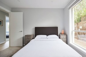 Ein Schlafzimmer mit einem großen Bett, einem hölzernen Kopfteil, zwei Nachttischen und einem Fenster mit Blick auf Grünfläche.