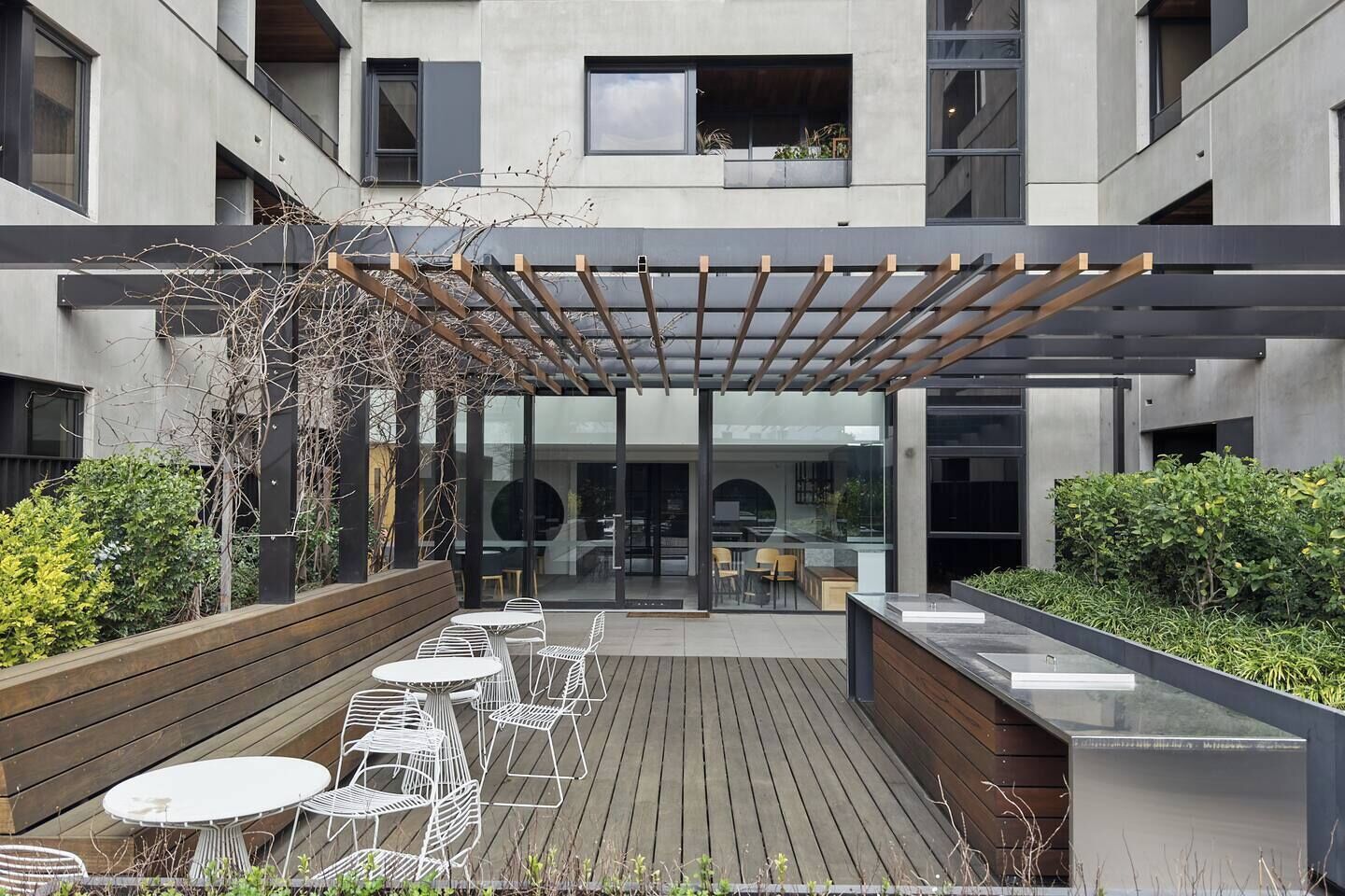 Een terras buiten met houten vloer, metalen tafels en stoelen, en een pergola.