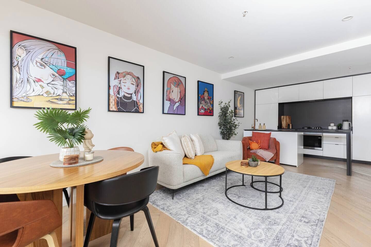Ein modernes Wohnzimmer mit einer Couch, einem Couchtisch und einem Essbereich. An der Wand hängen gerahmte Kunstwerke, und im Hintergrund ist eine Küche zu sehen.