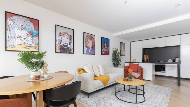 Uma sala de estar moderna com sofá, mesa de centro e área de jantar. Há quadros nas paredes e uma cozinha ao fundo.