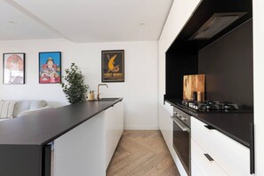 Una cuina moderna amb un taulell negre, armaris blancs, forn integrat i fogó. Hi ha quadres amb marc a la paret i una planta sobre el taulell.