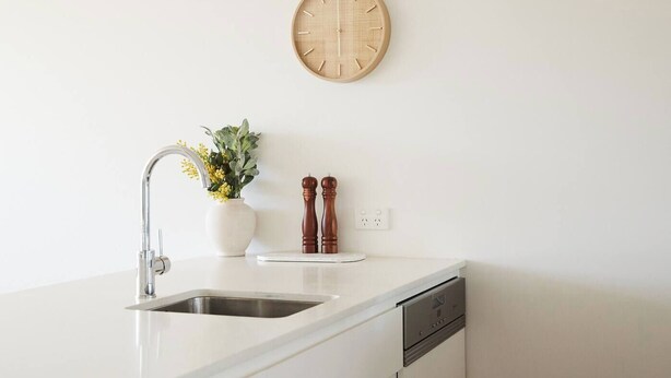 A kitchen with a white countertop, a stainless steel sink, a white vase with flowers, two wooden salt and pepper shakers, and a wooden clock on the wall. - Urban Drift Cosy Balcony Nook near Waterfront (Geelong)